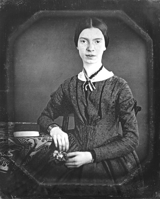 A young woman with dark hair parted in the center, seated with one hand resting on a table.