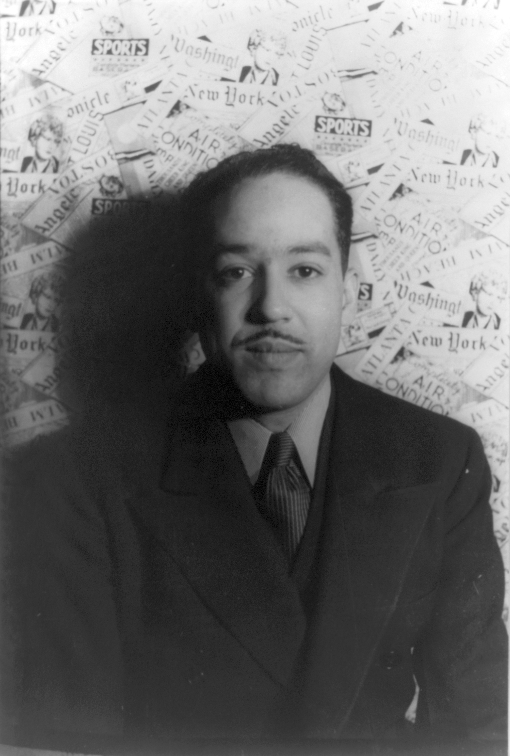A man in a suit and tie looks directly at the camera, resting his chin on his hand.