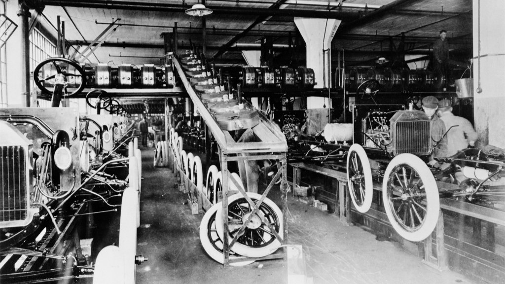 Ford Motor Company, Model T assembly line, Highland Park, Michigan, 1913. A long factory floor with rows of partially assembled automobiles on a moving production line, workers visible at their stations along both sides.