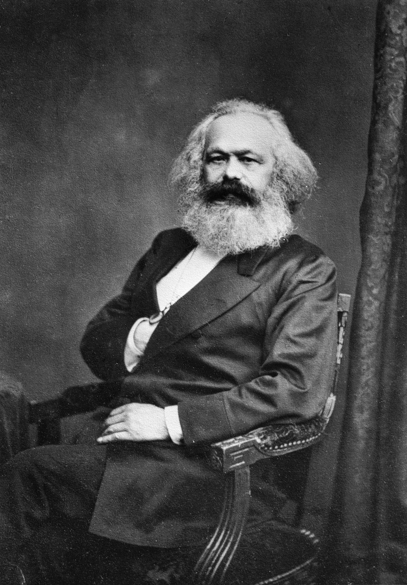 Portrait of Karl Marx, c. 1875. A bearded man in middle age looks directly at the camera, his expression composed and serious. He wears a dark jacket; his white shirt and cravat are visible at the collar.