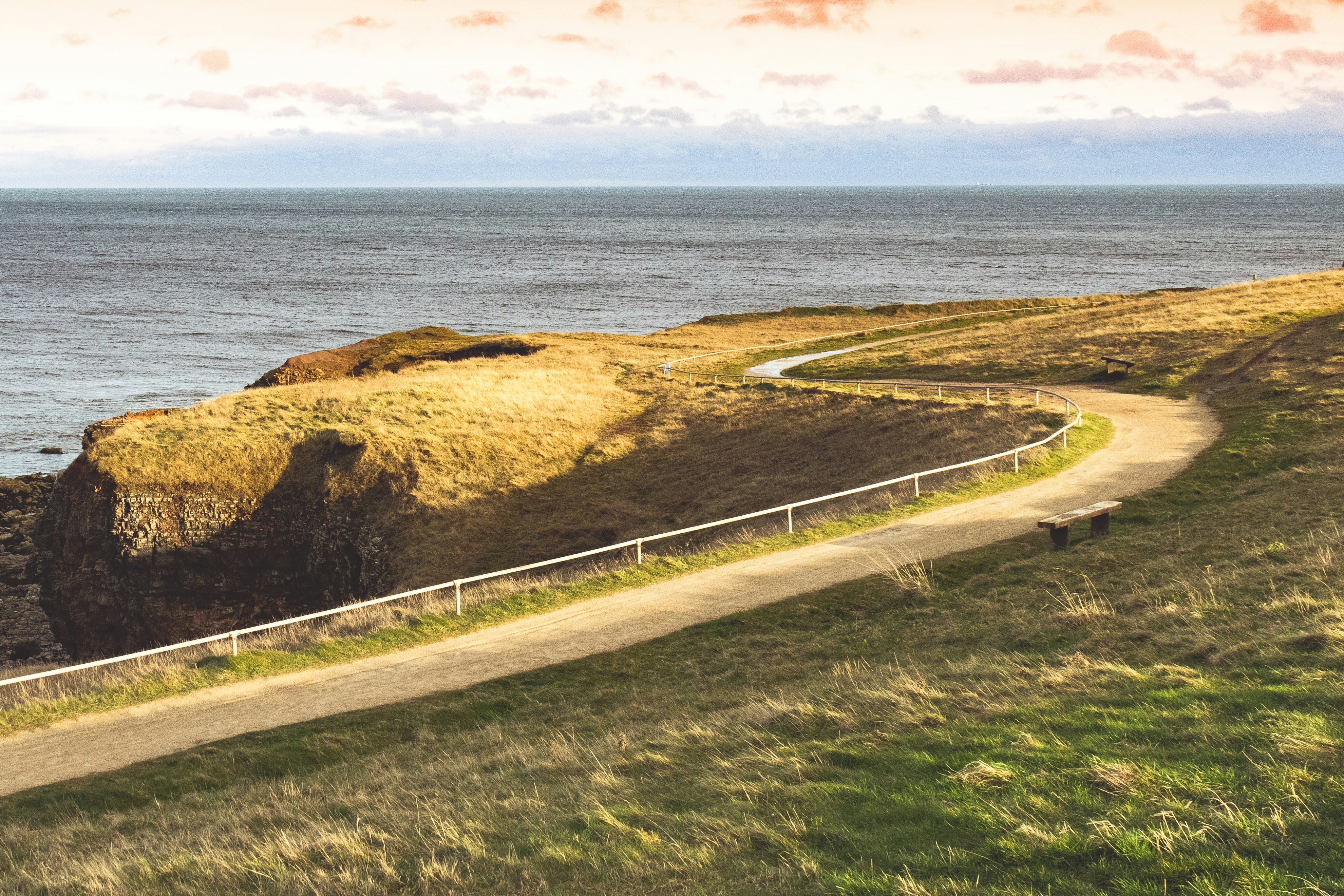 A coastal dirt road with a fence and ocean view during the day.